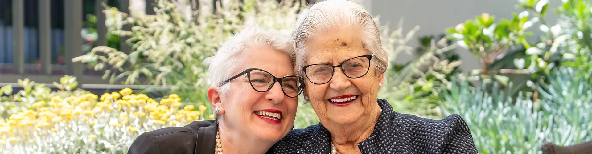 Two smiling women sitting at an outdoor table at Bert Newton Village in Highett, dressed in formal clothing and pearl necklaces, with flowers and greenery in the background.