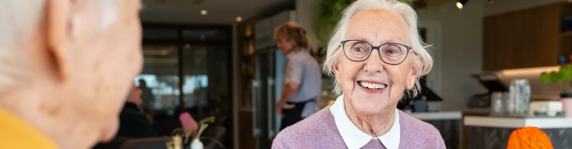 Smiling woman sitting at a table inside the Deborah Cheetham Village café, with another resident in the foreground and staff working in the background.