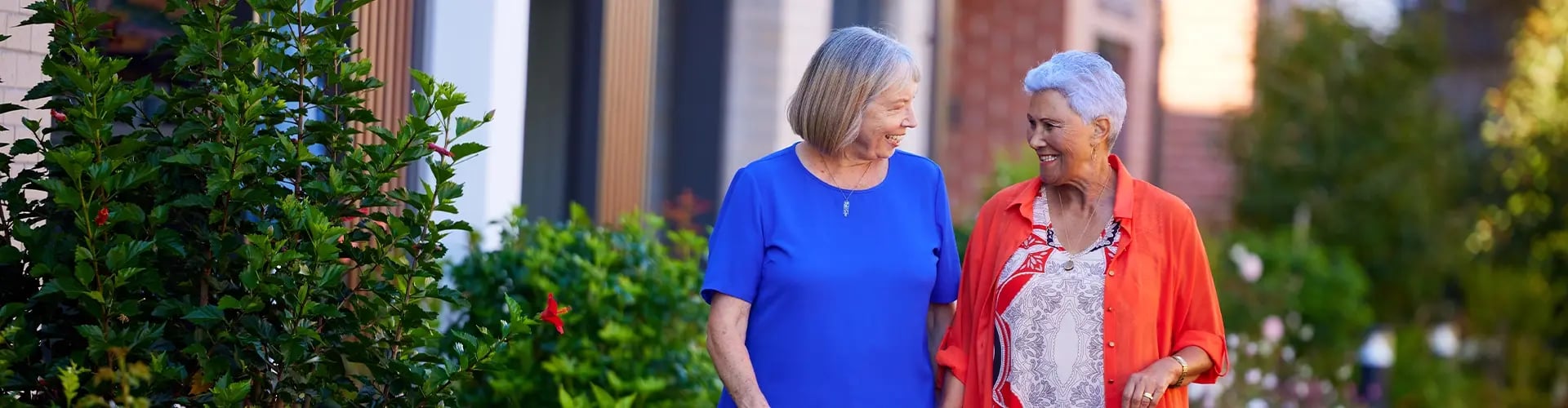 Two friends walking together along a garden-lined pathway at a Ryman retirement village, surrounded by flowers and greenery.