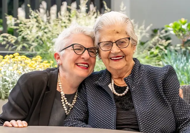 Two smiling women sitting at an outdoor table at Bert Newton Village in Highett, dressed in formal clothing and pearl necklaces, with flowers and greenery in the background.