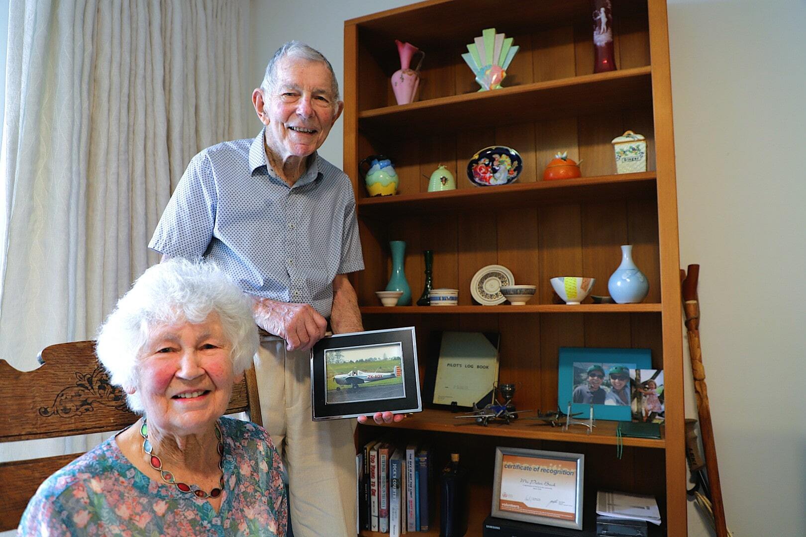 An older woman sits in a rocking chair next to an older man holding a photograph next to a bookshelf displaying precious objects from their life