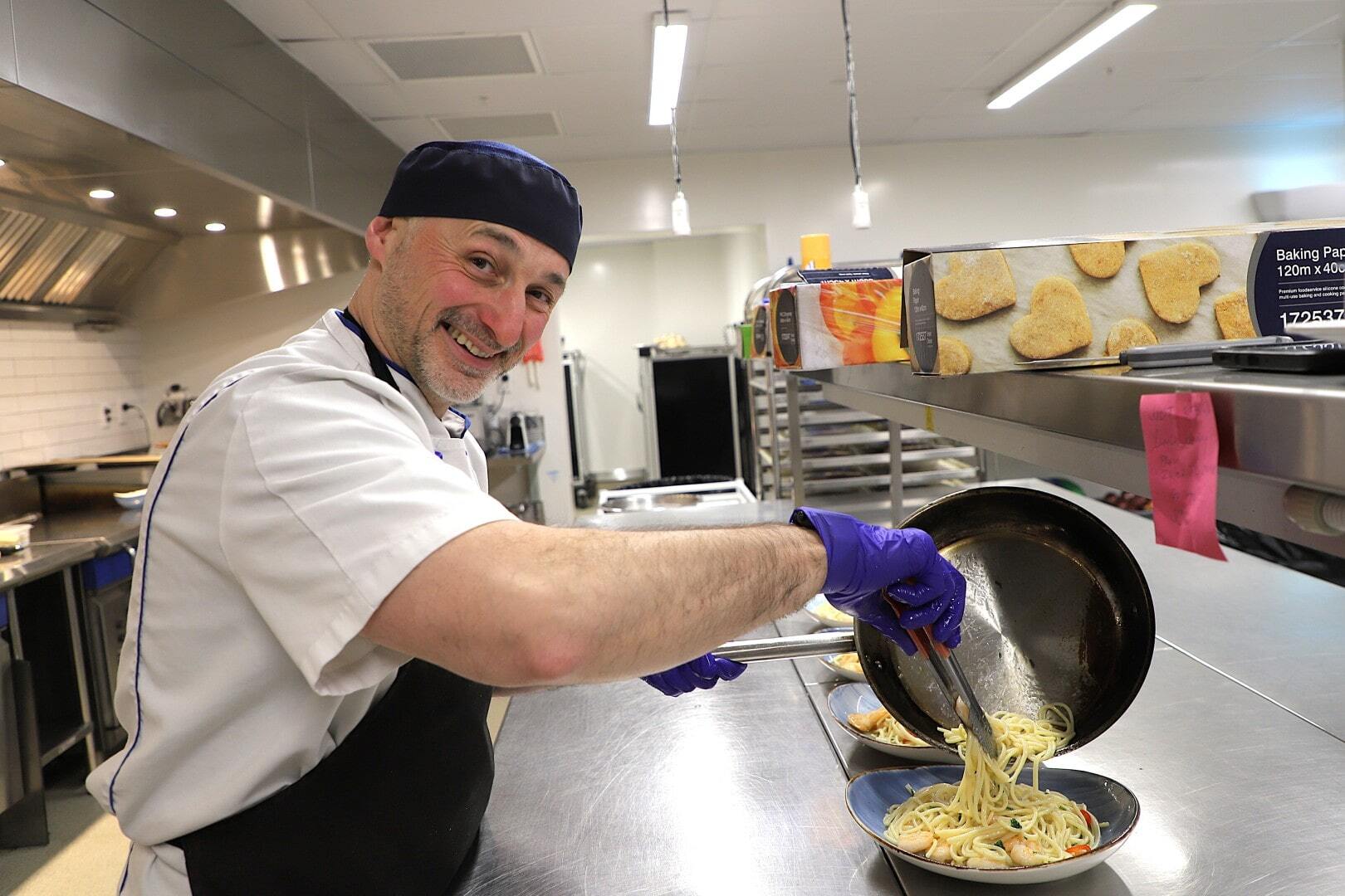 A smiling male chef in a commercial kitchen dishes up pasta from a frying pan into a bowl