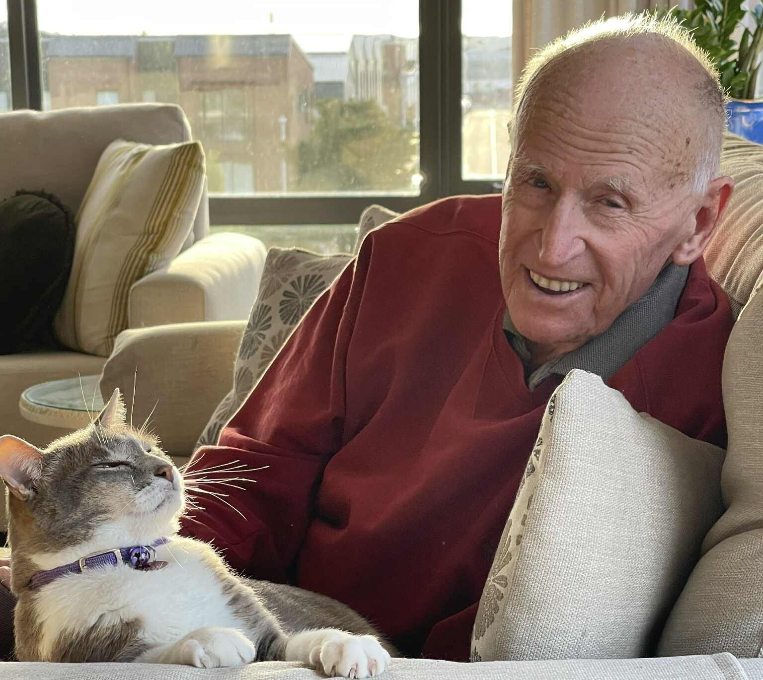 A smiling older man with his pet cat who sits next to him smiling